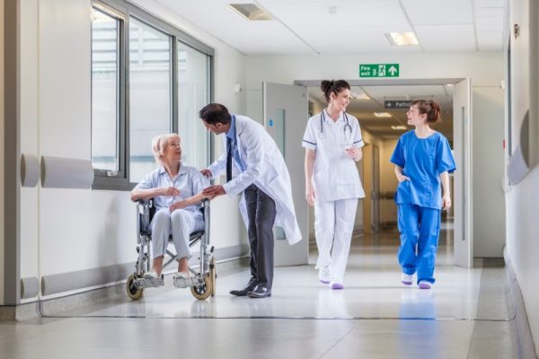 Senior female woman patient in wheelchair sitting in hospital corridor with Asian Indian male doctor and female nurse colleagues