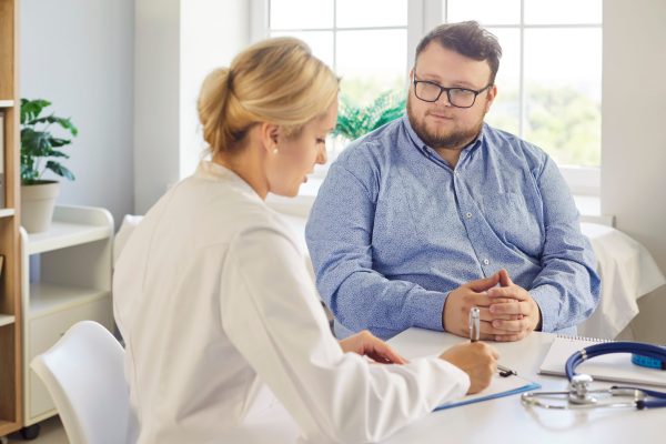 Physician writing notes while counseling obese patient during health consultation Doctor and patient during obesity consultation support. The physician writes notes as the fat overweight man listens during medical exam in clinic. Collaborative medical weight management